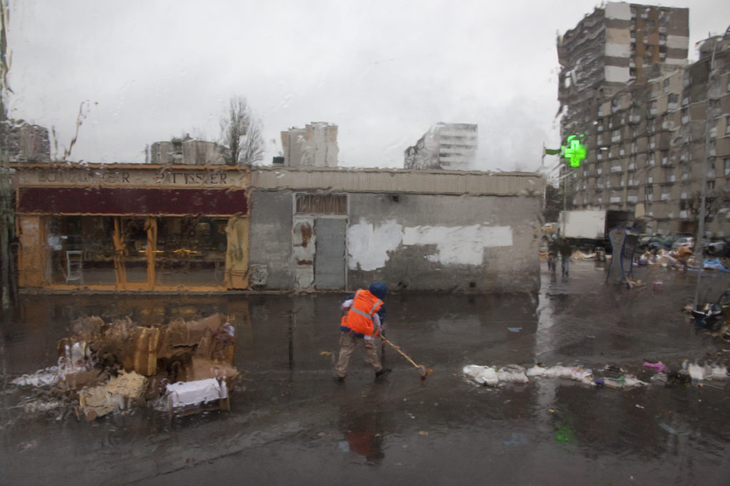 Stra§enkehrer kehrt hinter einer regennassen Scheibe die Reste eines Wochenmarktes zusammen/ cleaning the street in the rain after a local market